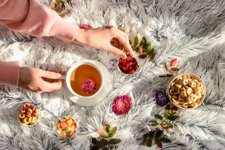 Woman with cup of hot tea, sweets, flowers and autumn leaves on fluffy fabric backgroundの写真素材