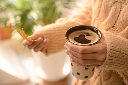 Woman with cup of hot coffee and autumn leaf at home, closeup