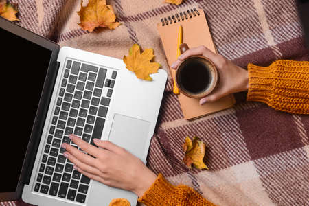 Woman with laptop, cup of coffee, notebook and autumn leaves on checkered fabric backgroundの写真素材