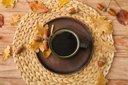 Wicker mat with cup of coffee, acorns and autumn leaves on wooden backgroundの写真素材