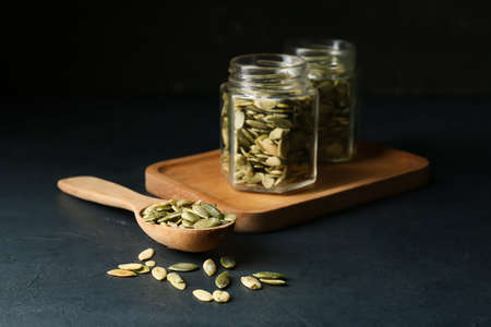 Spoon and jars with pumpkin seeds on black background
