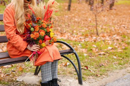 Young fashionable woman sitting on bench and holding bouquet in autumn parkの写真素材