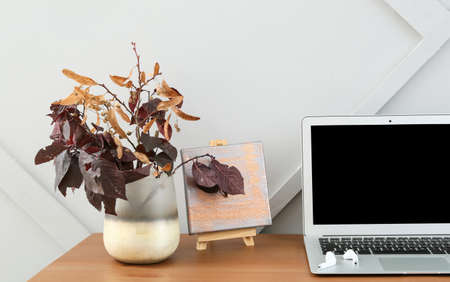 Modern laptop and vase with branches on table near light wallの写真素材