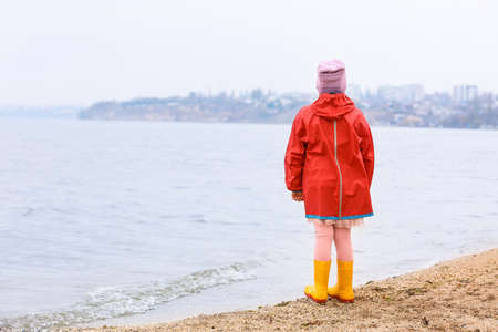 Little girl in stylish rubber boots and raincoat walking near the riverの写真素材