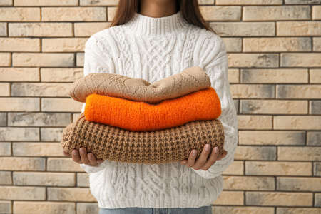 Young woman with beautiful manicure holding stack of warm knitted sweaters on brick wall backgroundの写真素材