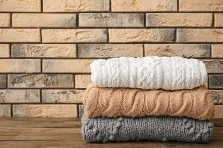Stack of stylish knitted sweaters on wooden table against brick wall background
