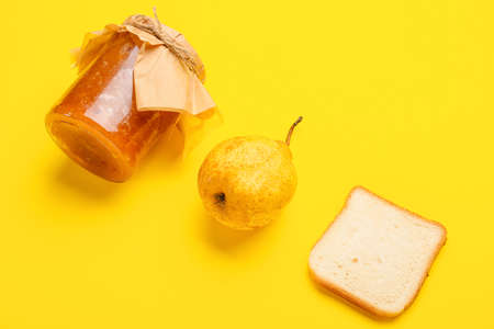 Glass jar of tasty pear jam and bread on yellow background