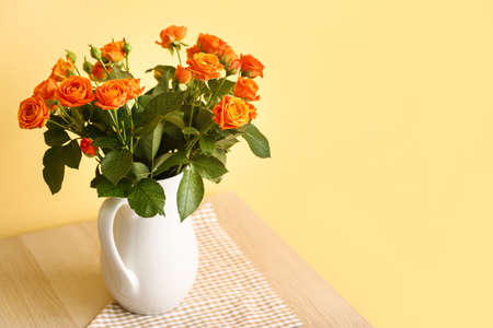 Jug with beautiful orange roses on table against yellow wall