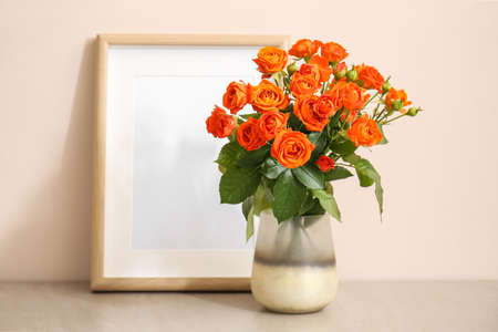 Vase with beautiful orange roses and blank photo frame on table against light wall