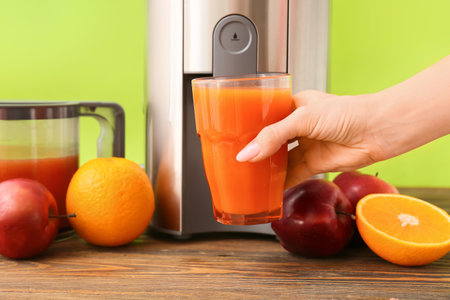 Female hand holding glass of fresh juice with fruits and new modern juicer on tableの写真素材