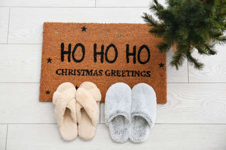Door mat with Christmas greeting, slippers and small fir tree on white wooden floorの写真素材