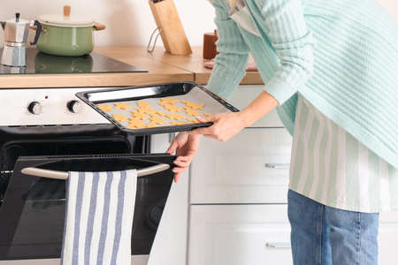 Mature woman putting baking dish with cookies into oven in kitchen