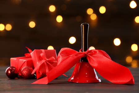 Christmas bell with ribbon and gift boxes on table against blurred lightsの写真素材