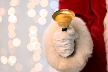 Santa Claus holding golden Christmas bell against blurred lights, closeup