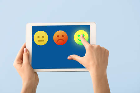 Female hands holding tablet computer and choosing emoticon on screen against light blue backgroundの写真素材