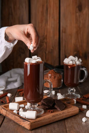 Woman adding marshmallows in glass cup with tasty hot chocolate on wooden background