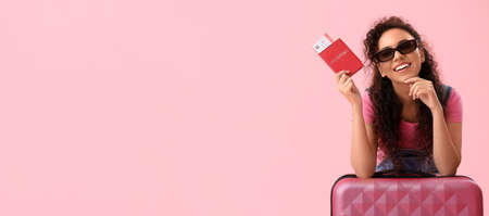 Young African-American woman with passport, tickets and suitcase on pink background with space for textの写真素材