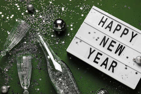 Board with text HAPPY NEW YEAR, champagne bottle, glasses and Christmas decor on green background,...