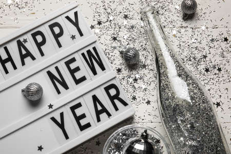 Board with text HAPPY NEW YEAR, champagne bottle, glass and Christmas decor on white wooden background, closeupの写真素材