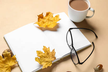 Cup of tasty coffee, blank book, autumn leaves and eyeglasses on beige backgroundの写真素材