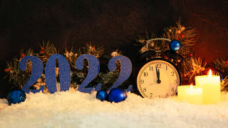 Composition with Christmas alarm clock, burning candles and figure 2022 on snow against dark...