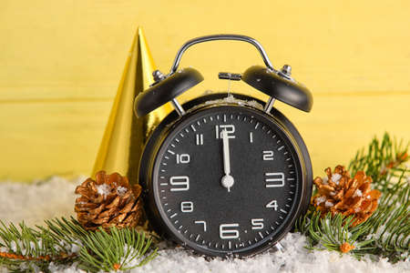 Christmas alarm clock, party hat, fir branches and pine cones on snow against color wooden...