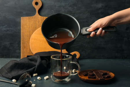 Woman pouring tasty hot chocolate from saucepan into glass cup on black background
