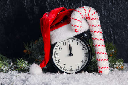 Composition with alarm clock, Santa hat and cane on snow against dark background