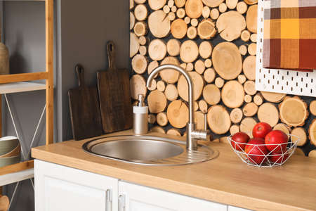 Wooden counter with silver sink, dish soap and basket of apples near wooden wall in kitchen