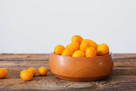 Bowl with tasty kumquat fruits on wooden table against light wallの写真素材