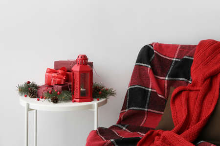 Red Christmas lantern with burning candle, gift boxes, decor on table and chair near light wallの写真素材
