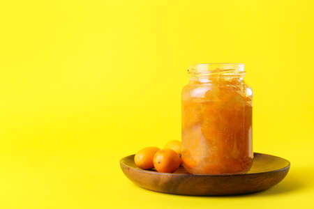 Jar of tasty kumquat jam and fresh fruits on yellow background