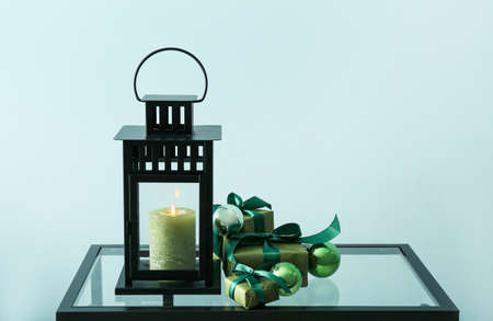 Lantern with burning candle, Christmas gifts and decorations on table near color wall