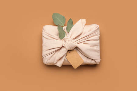 Gift box wrapped in cotton fabric with tag and eucalyptus branch on color backgroundの写真素材