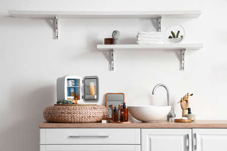 Open refrigerator with cosmetic products, mirror and sink on table top near light wall