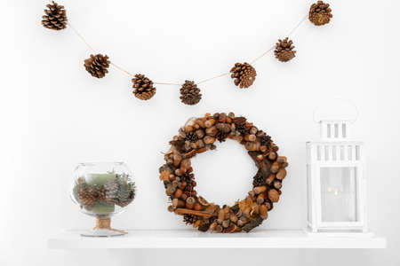 Shelf with pine cones, beautiful wreath and candle hanging on light wall in room