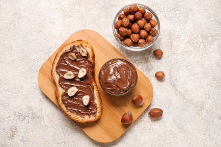Wooden board of bread with chocolate paste and hazelnuts on light background