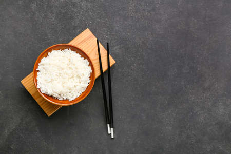 Bowl with tasty boiled rice and chopsticks on dark background