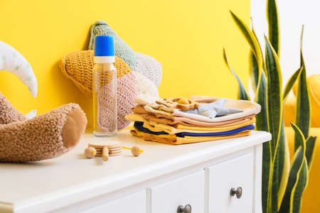 Stack of baby clothes with toys and bottle on table near yellow wall