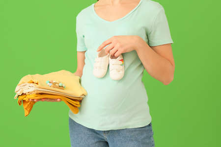 Pregnant woman with stack of baby clothes and boots on green background
