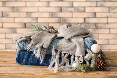 Knitted sweaters, scarf and fir tree branches on wooden table against brick wall background
