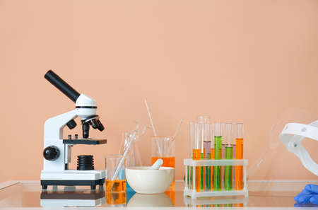 Modern microscope and laboratory glassware with liquid on table near beige wall