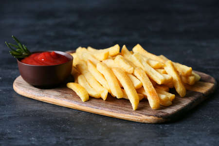 Wooden board with tasty french fries and ketchup on black background