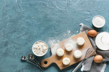 Wooden board with uncooked cottage cheese pancakes and ingredients on blue background