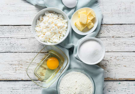 Bowl with cottage cheese and ingredients for preparing pancakes on white wooden background