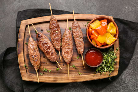 Wooden board of skewers with tasty lula kebab, sauce and grilled vegetables on black background