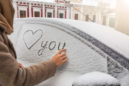 Woman drawing heart and word YOU on snow covered car outdoors