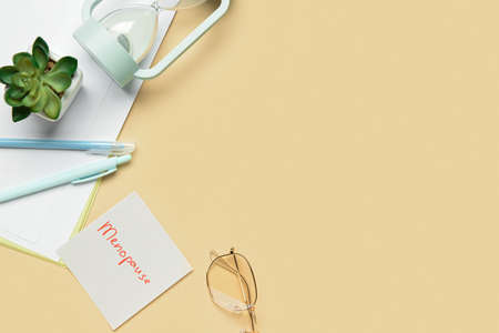 Sticky note with word MENOPAUSE, eyeglasses, pens, flowerpot and hourglass on beige background