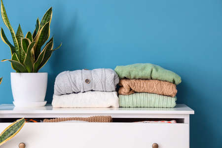 Stacks of sweaters and houseplant on chest of drawers near color wallの写真素材