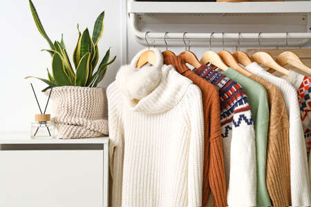 Hangers with stylish sweaters and ear muffs on rack near light wallの写真素材